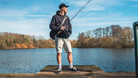 Hombre pescando en un muelle con pantalón corto gris Fox Rage Lightweight y chaqueta oscura, rodeado de naturaleza.