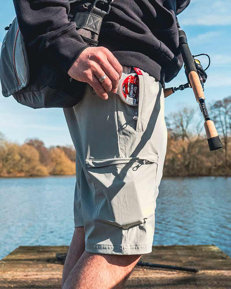 Hombre usando pantalón corto Fox Rage Lightweight gris mientras pesca en un lago, con caña y accesorios.